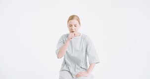 Camera zooming in and out at Caucasian young good looking woman in hospital night dress caughing as is ill with flu on white wall background. Female medical patient with coronavirus having bad caugh.