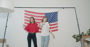 Two pretty stylish girls friends sisters celebrating independence day and having fun over national usa flag on white wall at home. 4k video footage slow motion