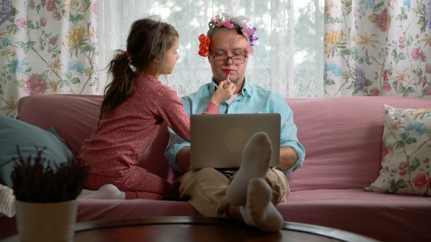 Home quarantine self-isolation. A middle-aged man working remotely from home. His little daughter getting in the way all the time. She tieing bows on his head, painting his lips and putting on makeup | Shutterstock HD Video #1050437104