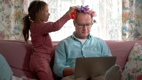 Home quarantine self-isolation. A middle-aged man working remotely from home. His little daughter getting in the way all the time and tieing bows on his head.