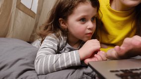 Side view of loving young mother teaching cute little daughter to use laptop, sitting in tent at home,  spend time together, mum and preschool child looking at electronic device screen