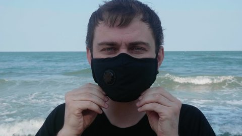 Smiling Travel Man Wearing Black Medical Face Mask, Looking at Camera and Showing Cool Gesture by Hands with Ocean on Background. Concept of Fight Against Chinese Covid-19 Coronavirus in Thailand