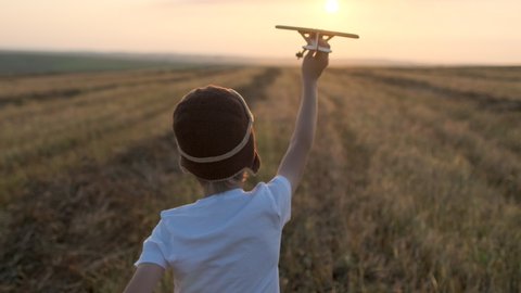 Happy little boy child in helmet aviator pilot of airplane running with toy wooden plane in field. Kid dreams of flying, traveling in summer nature at sunset outdoors. travel freedom dream 4 K slow-mo