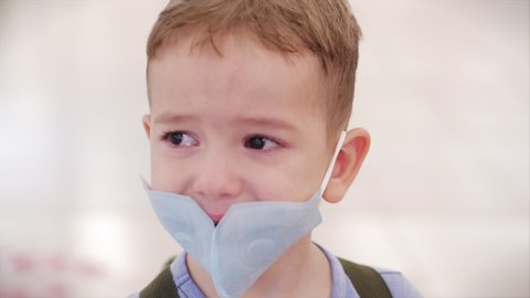 Portrait of a cute little boy a child in a mask from viruses of the coronavirus epidemic cries upset wipes eyes with his hands looks at the camera, tears crying frightened upset child looks around.
