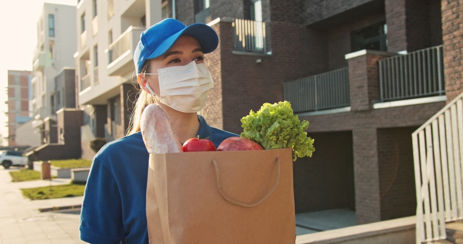 Caucasian young pretty woman, delivery worker in bluetooth headphones, cap and medical mask walking the street and carrying packet with fresh food. Female beautiful courier going to deliver grocery. Royalty-Free Stock Footage #1050465859