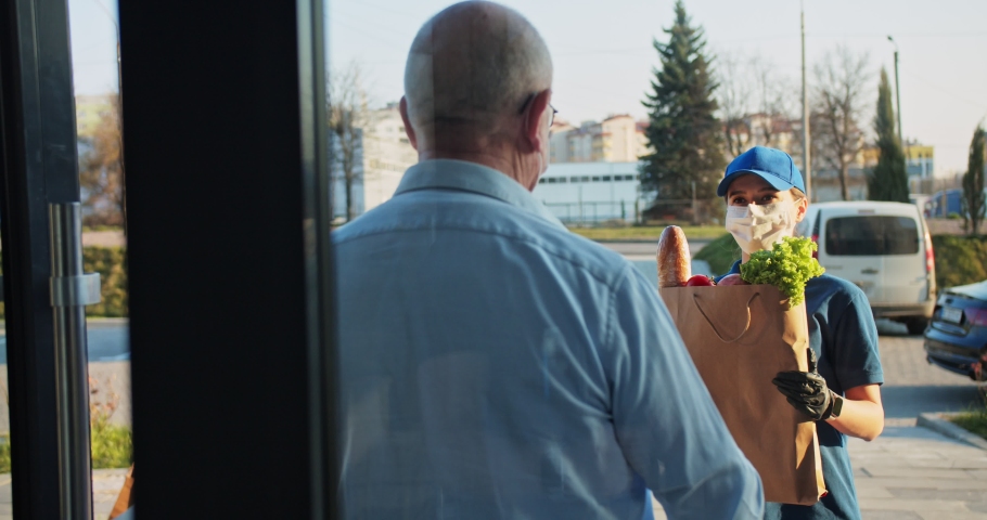 Rear senior man coming out from building and meeting young Caucasian female courier from supermarket. Woman in medical mask and gloves handing packet with food to male. Coronavirus isolation concept | Shutterstock HD Video #1050466042