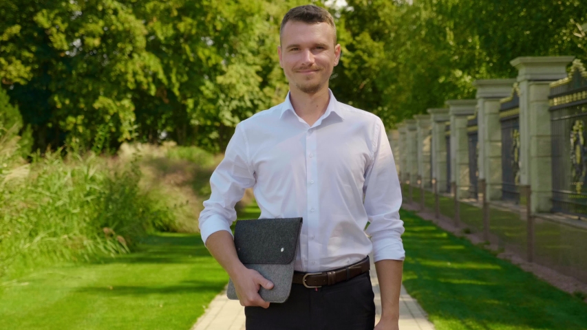 Businessman is standing in a park with a laptop in his hands, smiling and showing like. Freelancer man in a classic white shirt and trousers. 4K footage