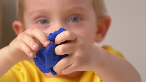 Child is sculpting with modeling clay. Portrait, close up face of little boy, hands crumpling blue compound (plasticine). Art activity for children, indoor fun for kids concept