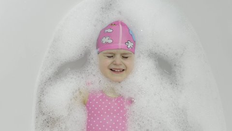 Attractive four years old girl takes a bath with bath foam in pink swimwear and swimming cap. Hygiene for cute blonde child. Cute girl smiling. Little blonde 4-5 year old child girl in bathroom