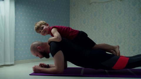 Bearded 30s father in sportswear making plank exercise with young son on his back. Happy family doing fitness together at home. 