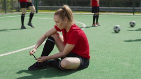 Female soccer player sitting on green playing field and tying shoelaces while her teammates exercising in the background - Powered by Shutterstock - Get 15% off with code: PIKWIZARD15