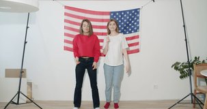 Two pretty stylish girls friends sisters celebrating independence day and having fun over national usa flag on white wall at home. 4k video footage slow motion