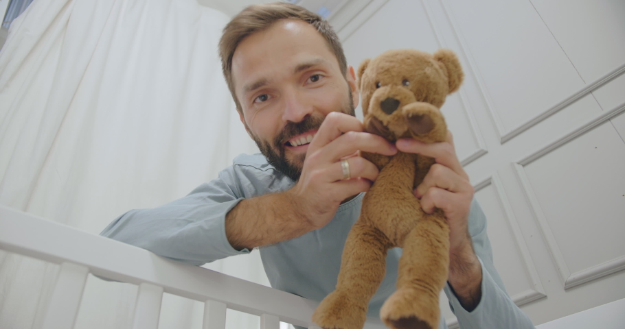 Low angle view of young happy father looking down and talking to newborn baby holding stuffed animal. Smiling parent enjoying time playing with infant in cradle. Royalty-Free Stock Footage #1050525052