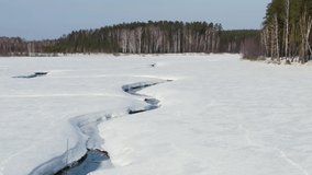 Flying in winter over a swamp covered with forest. A narrow, winding channel of dark open water cuts through a vast expanse of snow-covered ice, creating a stark linear contrast against the white frozen surface. The camera glides forward along the waterway, revealing the thick, layered ice edges that frame the dark liquid ribbon. On the right, a dense stand of tall, slender trees-mostly bare deciduous trunks mixed with dark evergreen pines-lines the frozen wetland, while a continuous forest wall stretches across the distant horizon under a pale, clear sky. The scene captures the quiet stillness of late winter or early spring in a boreal landscape, where the ice begins to yield to seasonal thaw. The aerial perspective emphasizes the scale of the frozen terrain and the meandering path of the waterway as it disappears into the flat, white distance. Dolly zoom. - Powered by Shutterstock - Get 15% off with code: PIKWIZARD15