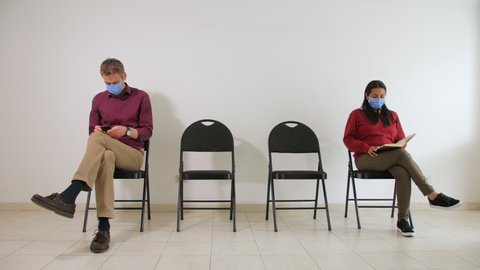 A man and a woman are sitting apart on chairs in a waiting room, keeping social distance, each with face mask, fearing covid-19 and coronavirus