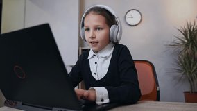 Front view of appealing satisfied 10-aged schoolgirl in earphones which sitting at the laptop, gaming and celebrating success after ending the video game
