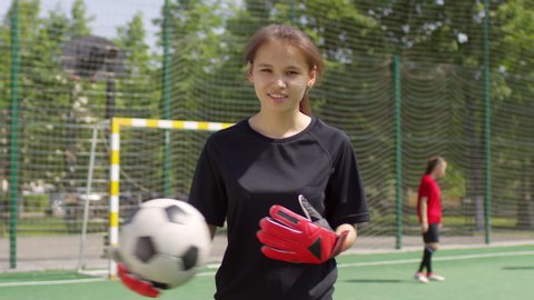 Young cheerful female goalkeeper in uniform and gloves holding soccer ball and looking at camera while standing on playing field outdoors