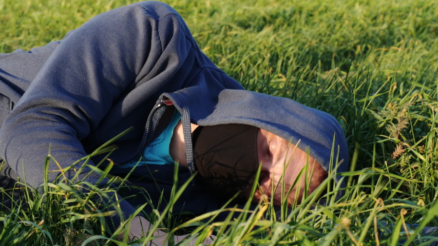 The Young Caucasian Man in Hoodie and Black Face Mask Lies in Green Field Outdoors. Concept of Unity with Nature, Healthy, Safety Life, Virus protection, Spreading Pandemic and World Panic Safety
