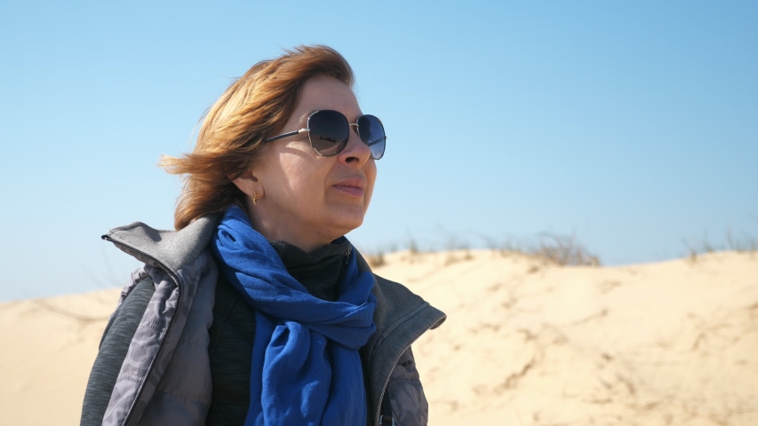 Wonderful view of a glamour fair-haired woman in sunglasses, fancy blue scarf, and a grey jacket looking at boundless sandy desert areas on a sunny day in spring