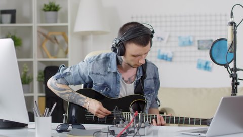 Young tattooed man in headphones playing the guitar and using console while recording music at home วิดีโอสต็อก