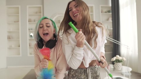 Two cheerful girls sing while cleaning the house. Friends have fun at home.