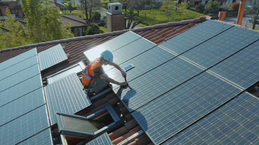 Aerial shot of worker with hard hat and safety equipment installing and working on maintenance of photovoltaic panel system installed on home domestic roof top, urban landscape. Wide angle camera. Royalty-Free Stock Footage #1050552082