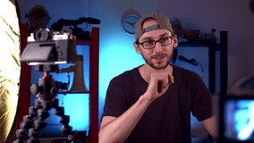 Caucasian bearded blogger with glasses and cap speaks video camera in studio, adjusts focus on the lens. Blog post for the internet. Blue background filming equipment. doll shot.