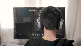 Boy playing in computer game. Teenager boy in gaming headphones sits in front of the monitor. The virtual reality. Kid in black hoodie with hood playing shooters or battle game in leaving room.