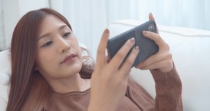 Portrait of Attractive Asian Woman using a smartphone playing a video game on mobile having fun and happy emotion lying on a couch in the living room at home. Relax and Leisure time concept.