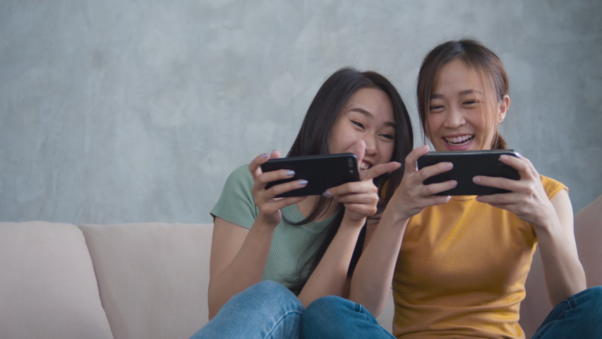 Portrait of Two female friends hand holing mobile phone sitting on a sofa playing a video game together with an excited and happy expression. Smiling woman having fun and enjoying in free time. Royalty-Free Stock Footage #1050586033