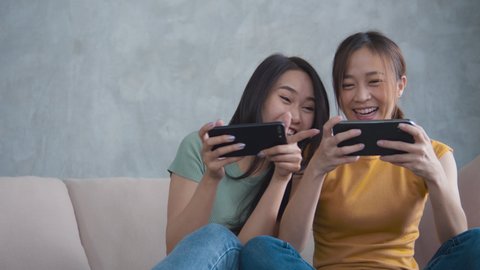 Portrait of Two female friends hand holing mobile phone sitting on a sofa playing a video game together with an excited and happy expression. Smiling woman having fun and enjoying in free time.