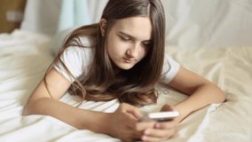 Happy relaxed teenage girl holding a smartphone using mobile applications, watching a funny video, laughing, lying on the bed, smiling, happily chatting on social networks, relaxing on the bed at home