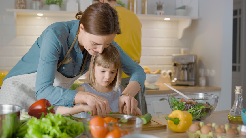 In the Kitchen: Mother and Cute Little Daughter Cooking Together Healthy Dinner. Mom Teaches Little Girl Healthy Habits and how to Cut Vegetables for the Salad. Cute Child Helping Her Caring Parents Royalty-Free Stock Footage #1050603106