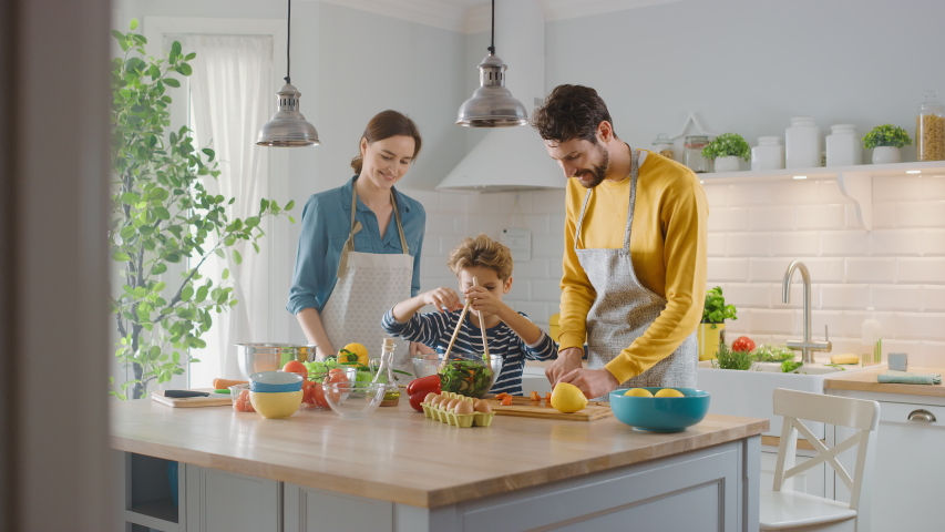 In the Kitchen: Mother, Father and Cute Little Boy Cooking Together Healthy Dinner. Parents Teach Little Son Healthy Habits and how to Mix Vegetables in the Salad Bowl. Cute Child Helping His Parents Royalty-Free Stock Footage #1050603109