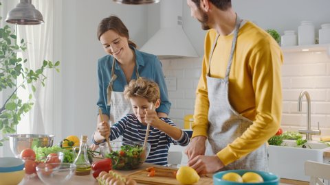 In the Kitchen: Mother, Father and Cute Little Boy Cooking Together Healthy Dinner. Parents Teach Little Son Healthy Habits and how to Mix Vegetables in the Salad Bowl. Cute Child Helping His Parents