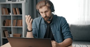 Mature businessman in headset talking during video conference on laptop while sitting at home. Bearded man in denim shirt working remotely during quarantine