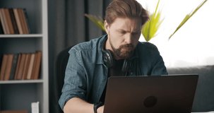Portrait of bearded man in denim shirt sitting at home and working on personal laptop. Concentrated mature person solving business issues online