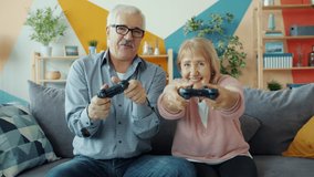 Slow motion of old people husband and wife enjoying videogame playing in apartment sitting on couch together. Modern technology, retirement and entertainment concept.