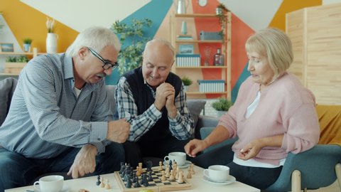 Elderly man and woman are playing chess while senior man friend is watching and laughing in apartment. Board games and entertainment concept.