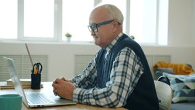Happy retired man enjoying online communication making video call with laptop talking gesturing looking at screen sitting at desk in modern apartment.