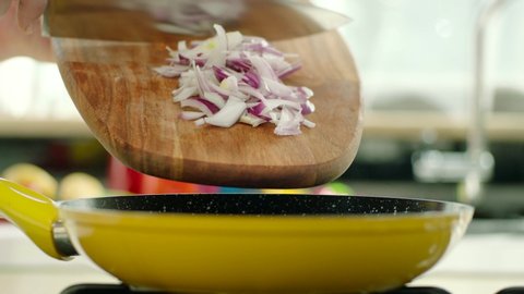 Chopped onions are thrown into the pan with a cutting board for cooking. Yellow pan on the stove, oil in the pan splashing. Slow motion.