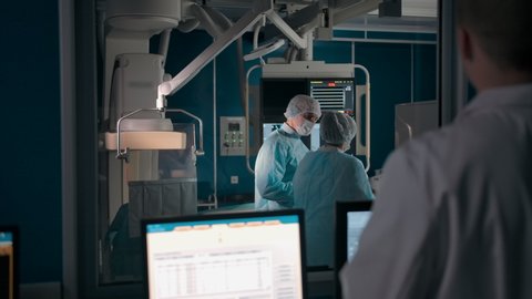 An experienced doctor oversees two fellow surgeons in the operating room. Monitors with patient disease data and X-rays in the observation room.