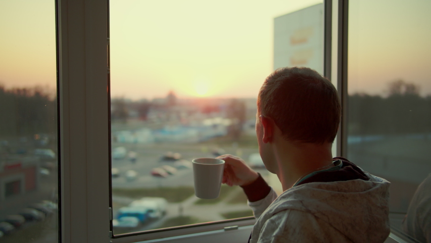A man watches the sunset and drinks tea from a white mug, rear view
