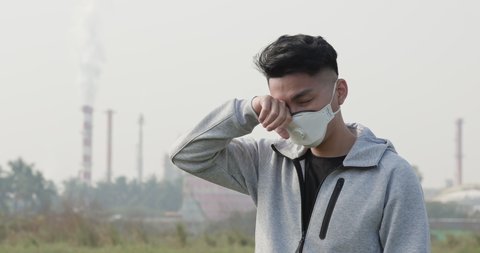 asian man wears protective n95 mask and has itchy eyes because air pollution in front of factory