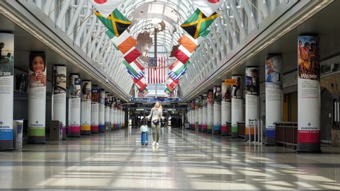 Chicago, IL / USA - April 16 2020: A young woman with a face mask around her neck walks down the eerily empty iconic hall of flags at O'Hare, through squares of sunlight, during the COVID-19 pandemic
