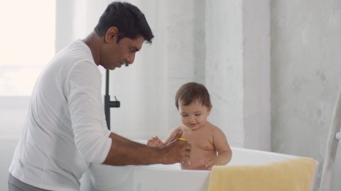 Handheld shot of cheerful Indian father smiling and giving yellow rubber duck and ball to happy toddler girl taking bath