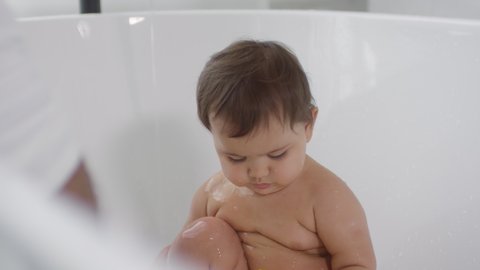 Close up shot of hands of unrecognizable father interacting and splashing water on curious toddler girl sitting in bathtub and playing with toys