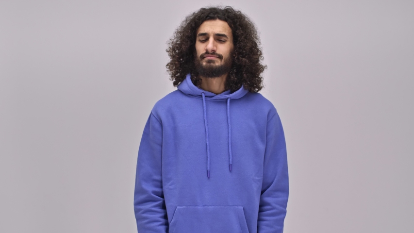 An irritated young bearded brunette man with long curly hair is driving a fly away isolated over the gray wall in the studio