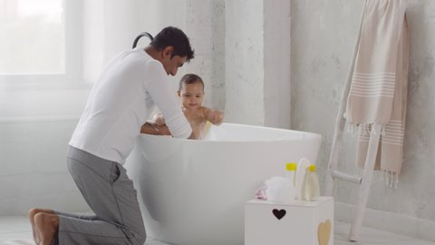 Medium shot of Indian father showing toys and interacting with cute toddler girl standing in bathtub during bath time
