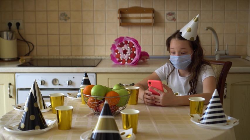 A girl in a festive cap and in a medical mask with a phone in her hands sits at the table alone on her birthday. Depression from lack of friends. Talking on a video call with friends. Coronavirus and  | Shutterstock HD Video #1050644287